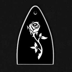 Puede incluir: Una p&uacute;a de guitarra negra con un dise&ntilde;o de rosa blanca. La p&uacute;a tiene forma de l&aacute;grima con un agujero en la parte superior. La rosa est&aacute; estilizada con un tallo y hojas.
