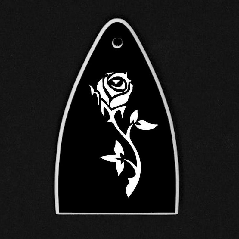 Puede incluir: Una p&uacute;a de guitarra negra con un dise&ntilde;o de rosa blanca. La p&uacute;a tiene forma de l&aacute;grima con un agujero en la parte superior. La rosa est&aacute; estilizada con un tallo y hojas.