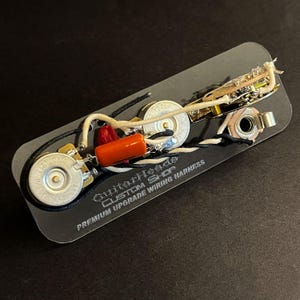 Può includere: Un cablaggio per chitarra rettangolare nero con vari componenti elettronici, tra cui potenziometri, un condensatore e cablaggio. Il testo "GuitarHeads CUSTOM SHOP PREMIUM UPGRADE WIRING HARNESS" è stampato sul cablaggio.