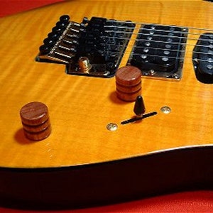 Puede incluir: Primer plano de una guitarra eléctrica marrón con un acabado de madera natural. La guitarra tiene dos botones de madera y un interruptor de palanca negro. La guitarra tiene una golpeador negro y un puente negro.