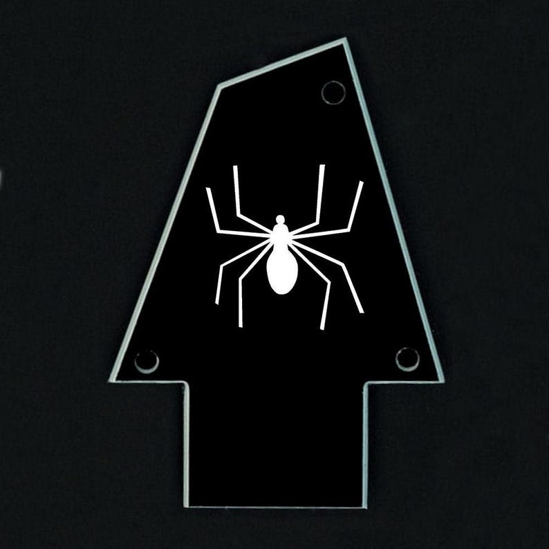 Pu&ograve; includere: Un oggetto nero a forma di freccia con una grafica di ragno bianco. Il ragno ha un corpo bianco e otto zampe bianche. L'oggetto ha tre piccoli fori e un bordo trasparente.