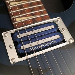 Könnte beinhalten: Nahaufnahme eines silbernen Gitarren-Tonabnehmers mit zwei blauen Humbucker-Tonabnehmern. Der Tonabnehmer ist mit "GuitarHeads" in einer stilisierten Schrift gekennzeichnet.