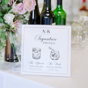 May include: A white sign with black text and illustrations of two cocktails. The sign reads "AK 10.08.2024 Signature Drinks". The groom's drink is called "Tom Collins" and the bride's drink is called "Tequila Smash".