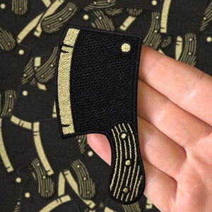 May include: A black embroidered patch shaped like a butcher knife with a gold outline and handle. The patch has a small circle on the blade.