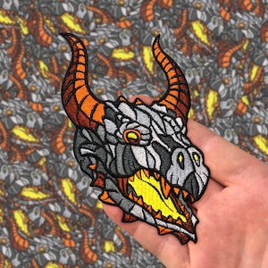 May include: A close-up of an embroidered iron-on patch featuring a dragon's head with orange and white scales, a yellow mouth, and black eyes. The patch is on a hand.