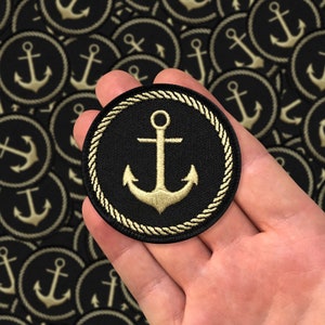 Könnte beinhalten: Ein schwarzer runder Patch mit einem Seilrand, auf dem ein goldener Anker gestickt ist. Der Patch wird in einer Hand gehalten.