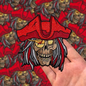 Könnte beinhalten: Gestickter Aufnäher mit einem Schädel, der einen roten Piratenhut mit einem weißen Augenklappen trägt. Der Schädel hat ein zahniges Grinsen und einen grauen Bart.