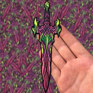 May include: A pink and green embroidered sword patch with intricate details. The patch is designed with a sharp point and a decorative handle.