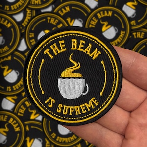 May include: Black and yellow embroidered patch with the text "THE BEAN IS SUPREME" surrounding a coffee cup graphic. The patch is round and has a yellow border. Several similar patches are visible in the background.