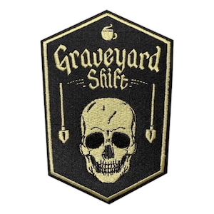 May include: Black and gold embroidered patch with the words "Graveyard Shift" above a skull and two shovels. A coffee cup is at the top. The patch has a gold border and is in a shield shape.