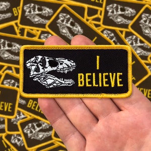 May include: A black and yellow embroidered patch with a white dinosaur skull and the text "I BELIEVE".