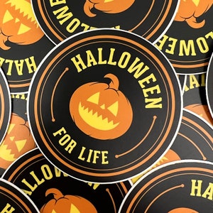 May include: A black and orange round sticker with the text "Halloween for Life" in a yellow font. A cartoon orange pumpkin with a yellow face is in the centre of the sticker.