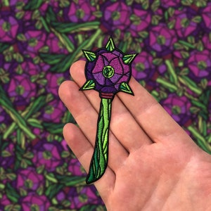 Puede incluir: Un parche bordado que representa una maza con púas con un diseño floral. La maza es de color morado y verde, con un tallo y hojas verdes.