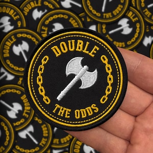 Op de afbeelding: Een geborduurde zwarte en witte patch met een witte bijl en een gouden kettingontwerp. De tekst "DOUBLE THE ODDS" is in geel op de patch geborduurd.