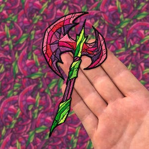 May include: A pink and green embroidered axe patch with a jagged blade and a detailed handle. The patch is held in a hand.