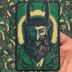 May include: Green and black embroidered patch featuring a horned, bearded figure with a menacing expression. The figure is surrounded by a swirling, vine-like pattern.
