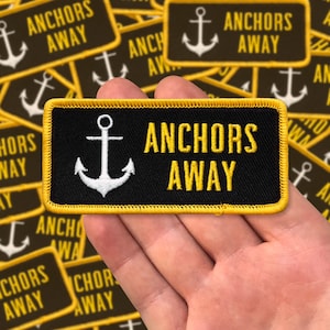 May include: A black embroidered patch with yellow stitching. The patch features a white anchor and the text "Anchors Away" in yellow thread.
