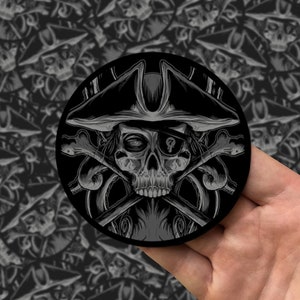 Puede incluir: Una ilustración en blanco y negro de una calavera con un sombrero de pirata y un parche en el ojo. La calavera sostiene dos huesos cruzados. La ilustración tiene forma circular.