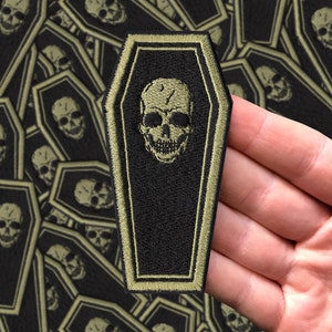 May include: A black and green embroidered patch shaped like a coffin with a white skull inside. The patch has a green border.