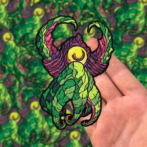 May include: Embroidered patch featuring a stylized creature with green, purple, and pink details. The design includes a large yellow eye and intricate patterns, perfect for jackets, bags, or other fabric items.