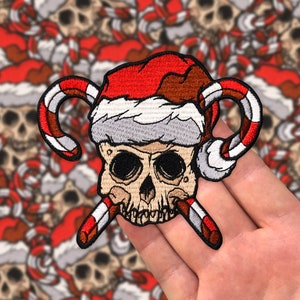 May include: A red and white candy cane skull wearing a Santa hat. The skull is embroidered with white and black details. The candy canes are crossed behind the skull.