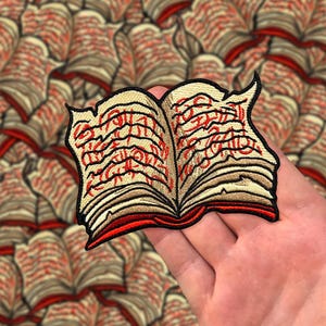 May include: A red and beige embroidered patch of an open book with a detailed design of swirling text. The book is open and the pages are curled.
