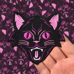 Peut inclure: Patch brodé noir à repasser représentant un visage de chat avec des oreilles, des yeux et une bouche roses. Le chat grogne avec sa langue qui sort.