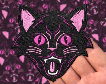 May include: Black embroidered iron-on patch of a cat's face with pink ears, eyes, and mouth. The cat is snarling with its tongue sticking out.