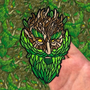 May include: Embroidered patch of a green man with a brown face, green beard, and leaves for hair. The eyes are yellow and the mouth is a small red line.