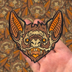 May include: A brown and orange embroidered patch of a bat's face with sharp teeth and glowing yellow eyes.