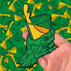 May include: A green monster hand holding a green glass with a yellow liquid inside. The hand is holding the glass up to the viewer. The background is a green and yellow pattern.