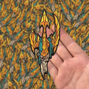 May include: Embroidered patch featuring a stylized, golden trident design with teal and orange accents. The patch has a detailed, textured appearance, with a cream-colored base. The patch is held in a hand against a patterned background.