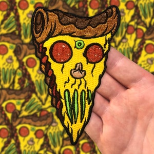 Puede incluir: Una rebanada de pizza amarilla con pepperoni y pimientos verdes, bordada con una cara de calavera y una corteza marrón. La rebanada de pizza es un parche.