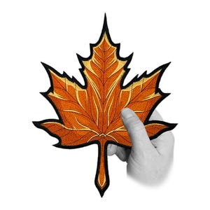 Pode incluir: Remendo bordado de folha de bordo em tons de laranja, dourado e preto. A folha é segurada por uma mão contra um fundo branco. O remendo apresenta costuras detalhadas e um design clássico de outono.