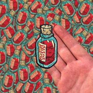 Puede incluir: Parche bordado con una botella de poción turquesa con tapón de corcho. La botella contiene una sustancia roja y una etiqueta con la palabra "DREAMS". El parche tiene un borde negro y está sobre un fondo de parches similares.