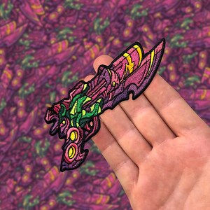Peut inclure: Un patch brodé représentant une arme futuriste en rose, vert et jaune. Le patch est finement conçu et est tenu dans une main.