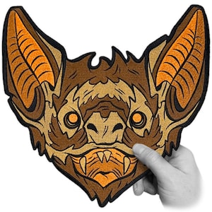 Op de afbeelding: Een geborduurde patch met het gezicht van een vleermuis, in de kleuren bruin en oranje, met scherpe tanden en grote oren.
