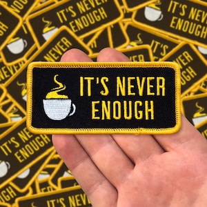 Puede incluir: Un parche rectangular negro con ribete amarillo y el texto "IT'S NEVER ENOUGH" en amarillo. Una taza de café blanca con vapor amarillo está a la izquierda. El parche se sostiene en una mano, con otros parches en el fondo.