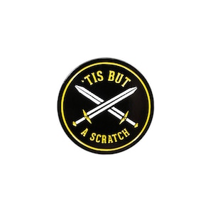 Könnte beinhalten: Ein runder, schwarzer Anstecker mit gelbem Rand und dem Text "'TIS BUT A SCRATCH" in Gelb. Zwei gekreuzte weiße Schwerter befinden sich in der Mitte. Der Hintergrund ist weiß.