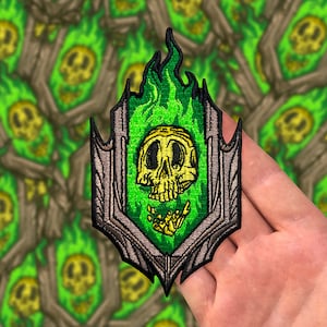 Könnte beinhalten: Grün-grauer gestickter Aufnäher mit einem Totenkopf, hinter dem sich eine grüne Flamme befindet. Der Totenkopf ist gelb und hat ein detailliertes Design. Der Aufnäher ist von einem dekorativen Rahmen umgeben.