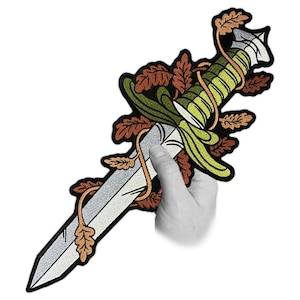 May include: Embroidered patch featuring a dagger design with a silver blade, green and yellow handle, and brown and orange leaves. The patch is held by a hand against a white background. The patch is approximately 20 cm long.