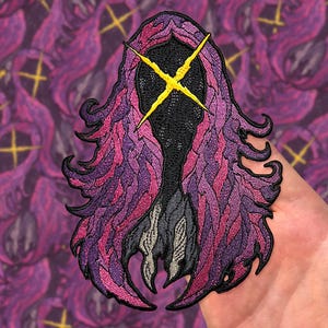 Op de afbeelding: Geborduurde patch met een gestileerde figuur met een zwart gezicht en een geel kruis. De figuur heeft vloeiende paarse en roze haarachtige details. De patch is bedoeld voor kleding of accessoires.