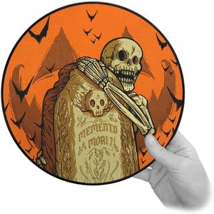 Könnte beinhalten: Runder Aufnäher mit Halloween-Motiv. Er zeigt ein Skelett, das einen Grabstein mit der Aufschrift "MEMENTO MORI" umarmt. Der Hintergrund ist orange mit Fledermäusen und Bergen.
