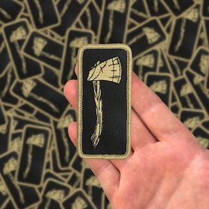 May include: A black and tan embroidered patch with a stylized axe design. The patch has a tan border and is held in a hand.