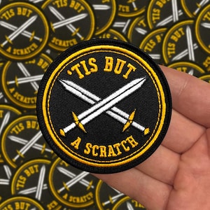 Könnte beinhalten: Runder schwarzer Aufnäher mit gelbem Rand und dem Text "'TIS BUT A SCRATCH". Zeigt zwei gekreuzte weiße Schwerter mit gelben Griffen. Weitere Aufnäher sind im Hintergrund zu sehen.