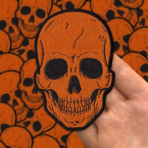 Puede incluir: Parche de calavera bordado de color naranja con costuras negras. La calavera tiene un diseño detallado con una mandíbula y dientes prominentes.