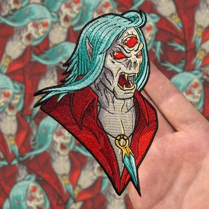 Puede incluir: Parche bordado que representa un vampiro estilizado con cabello turquesa, ojos rojos y un abrigo rojo. El parche tiene un diseño detallado con un colgante dorado y una daga azul. El fondo muestra un patrón de imágenes de vampiros similares.