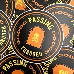 Peut inclure: Un autocollant rond noir et orange avec le texte "Passing Through" en lettres jaunes. L'autocollant représente un crâne et des os croisés à l'intérieur d'une arche en forme de pierre tombale avec une bordure de chaîne.