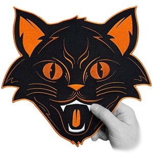 Peut inclure: Patch Halloween visage de chat noir avec des détails brodés orange. Le chat a la bouche grande ouverte avec une dent blanche et une langue orange.