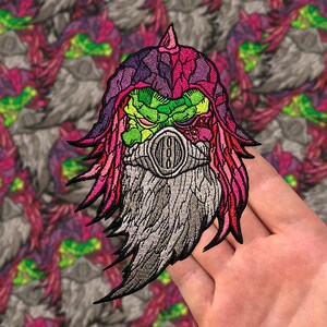 May include: A pink, purple, and green embroidered patch of a creature with a gas mask. The creature has a long, flowing beard and a menacing expression.